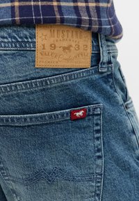 Jeans denim Mustang w jasnoniebieskim odcieniu, z brązową skórzaną metką i czerwoną łatą z logo na tylnej kieszeni. Widać kratkowany materiał powyżej.