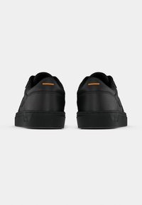 Svarte atletiske sko med glatt skinnstruktur, som har en gummisåle, avrundet design og en liten oransje detalj på hælen.