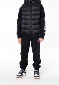 Černá prošívaná vesta s kapucí, se zapínáním na zip, nošená přes černou mikinu a kalhoty. Na rukávu je logo Champion a na nohou má tenisky v námořnické modři, bílé a oranžové barvě.