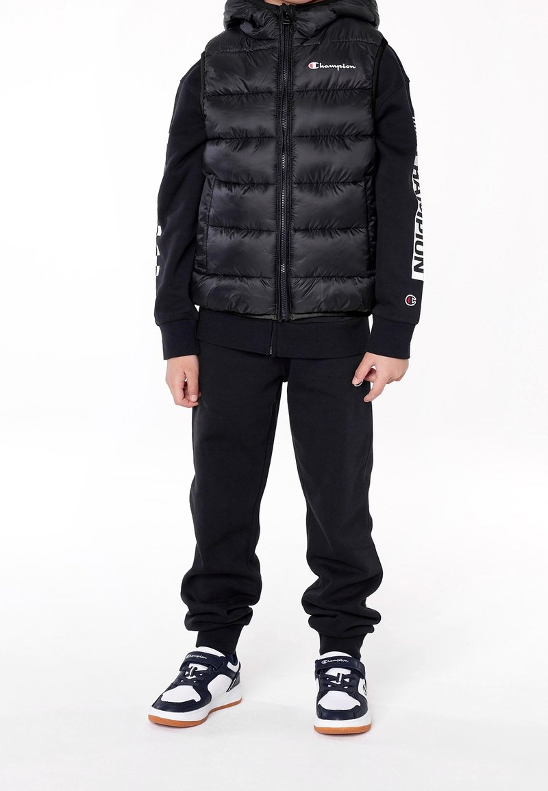 Černá prošívaná vesta s kapucí, se zapínáním na zip, nošená přes černou mikinu a kalhoty. Na rukávu je logo Champion a na nohou má tenisky v námořnické modři, bílé a oranžové barvě.