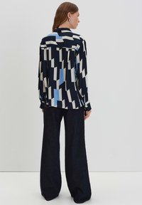 Camicia a maniche lunghe di colore blu navy con motivi geometrici in azzurro chiaro, crema e nero. Presenta un colletto e polsini con bottone. Indossata con jeans scuri a gamba larga.