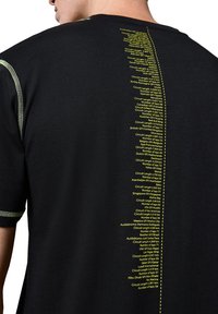 Czarna koszulka z krótkim rękawem z zielonymi akcentami krawieckimi. Z tyłu znajduje się żółty tekst z różnymi szczegółami toru wyścigowego, umieszczony pionowo.