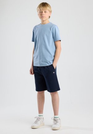 T-shirt di cotone azzurro chiaro, pantaloni corti blu navy con tasche e dettagli del logo, sneakers bianche con accenti grigi e calze alla caviglia a righe verdi.