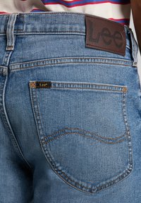 Vaaleansinisen farkkujen takataskussa on ruskea nahkainen etiketti, johon on painettu "Lee", ja oranssit tikit koristelevat taskua.