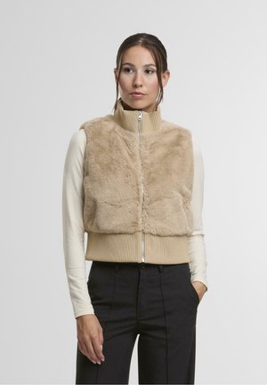Gilet in pelliccia sintetica beige con colletto alto, chiusura con zip e orlo a coste. Indossato sopra una maglietta a maniche lunghe leggera, abbinato a pantaloni scuri.