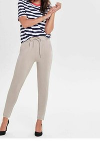 Gestreiftes navyblaues und weißes T-Shirt mit kurzen Ärmeln. Hellbeige, figurbetonte Hose mit Kordelzug. Schwarze High Heels. Schlichter Hintergrund.