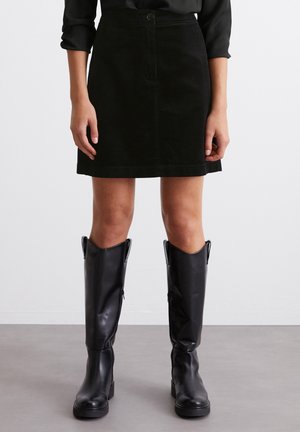 Vrouw die een zwarte ribfluwelen minirok met knopen aan de voorkant draagt en zwarte leren laarzen tot over de knie, staand op een grijze vloer.