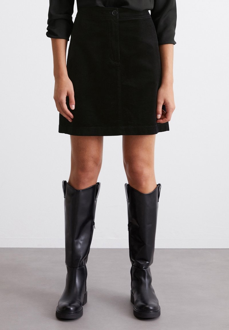 Femme portant une mini-jupe en velours côtelé noir avec boutons sur le devant et des bottes en cuir noires montant jusqu'aux genoux, debout sur un sol gris.