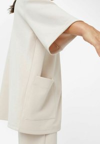 Beige oversized top s kulatým výstřihem a širokými krátkými rukávy, s boční kapsou. Měkká textura a minimalistický design.