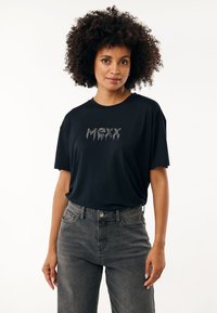 Schwarzes T-Shirt mit kurzen Ärmeln und lockerer Passform, mit metallic Textdesign "MAXX" auf der Brust. Kombiniert mit hoch taillierten grauen Jeans.