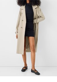 Beige trenchcoat med dubbelknäppning, bärs över en svart figurnära klänning. Svarta vävda ballerina-skor fulländar outfiten och framhäver en avslappnad look.