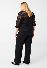 Zwarte kant blouse met driekwart mouwen en textuurpatronen, gecombineerd met zwarte wijde broek en platte zwarte schoenen.