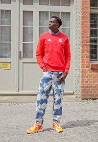 Rød Adidas hettegenser med tre striper, blå bukser med sky-mønster, og oransje sportssko. Står foran en dør.