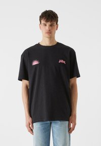 Must mustac t-särk, millel on roosa päike ja tekstikujundused rinnal. Üks suurus suurem istuvus lühikeste varrukatega ja ümmargune kaelus.