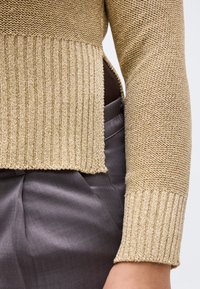 Pull en maille beige avec ourlet et poignets côtelés, détail fente latérale, porté sur un pantalon gris taille haute.