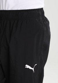 Svarta Puma-träningsbyxor med elastisk midja, bärs med vit skjorta, visar den vita Puma-logotypen på vänster lår.