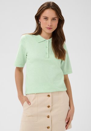 Mladá žena s dlhými hnedými vlasmi má na sebe mentolovo zelené polo s krátkym rukávom a béžovú sukňu so zapínaním na gombíky vpredu, stojí s jednou rukou v vrecku.