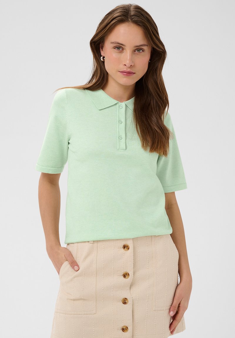 Jeune femme aux longs cheveux bruns portant un polo manches courtes vert menthe et une jupe beige à boutons sur le devant, debout avec une main dans la poche.