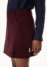 Burgunderfarbene Cordminirock mit gerippter Textur, seitlichem Schlitz und kleinem Markenetikett am Saum. Getragen mit einer marineblauen Langarm-Button-up-Hemd.