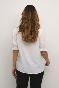 Blusa bianca con maniche corte a palloncino e polsini arricciati, caratterizzata da una texture liscia e una vestibilità rilassata, abbinata a pantaloni scuri.