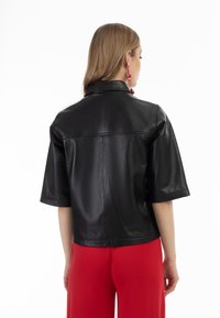 Jachetă din piele neagră cu mâneci scurte și guler, având un design curat și o textură netedă, purtată peste pantaloni roșii.