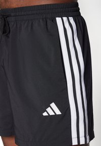 Shorts de sport noirs en tissu léger avec trois bandes verticales blanches sur les côtés et un logo blanc sur la jambe.