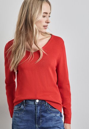 Roter Strickpullover mit V-Ausschnitt und gerippten Bündchen, mit einer leicht taillierten Silhouette. Kombiniert mit dunkelblauen Jeans.
