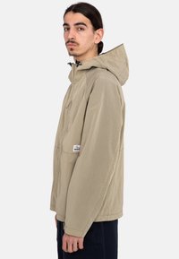 Chaqueta a cuadros de color marrón claro con capucha, que cuenta con un bolsillo frontal y puños ajustables. El material parece texturizado y resistente a las inclemencias del tiempo.