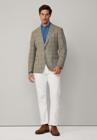 Blazer marrone a scacchi in un tessuto strutturato, camicia blu sotto, pantaloni bianchi e scarpe marroni stringate. Sfondo neutro.