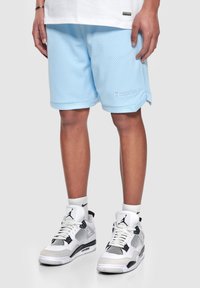 Pantalones cortos de malla de color azul claro con un logo bordado en el dobladillo, combinados con zapatillas deportivas blancas y negras y calcetines blancos.