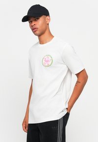 T-shirt de algodão branca com decote redondo e mangas curtas, apresentando um design gráfico colorido no peito. Boné preto e calças de ganga escuras.