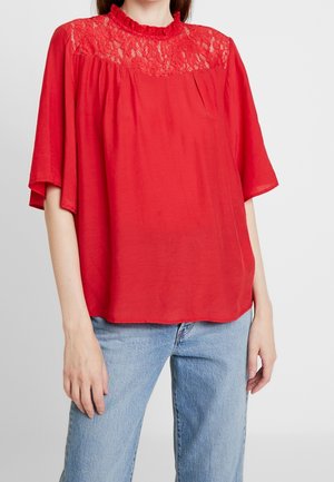 Haut rouge avec empiècement en dentelle, manches courtes évasées et encolure froncée. Associé à un jean bleu clair. Tissu lisse à texture légère.