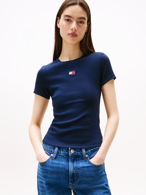 Camiseta ajustada de color azul marino con mangas cortas y textura acanalada, con un pequeño parche de logo en el pecho. Combinada con jeans de color azul claro.