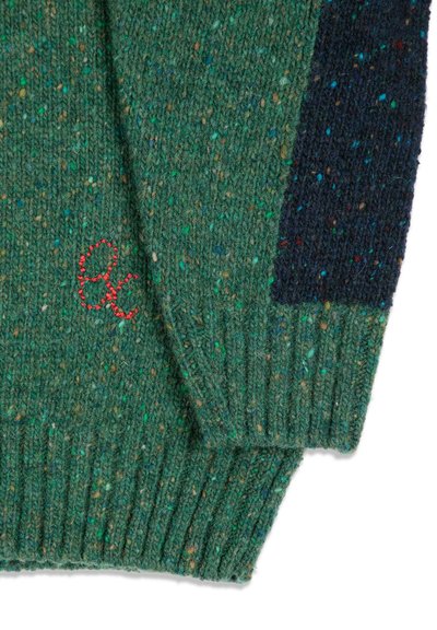 Gros plan d'une manche de pull tricoté vert et bleu marine moucheté avec des lettres minuscules rouges brodées « "ac" » près du poignet.