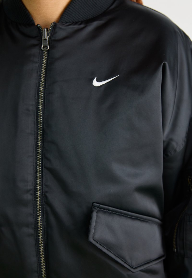 Veste bomber noire avec une texture lisse et brillante. Dotée d'une fermeture éclair à l'avant, d'un col à côtes et d'un logo Nike blanc sur la poitrine gauche. Comprend une poche.