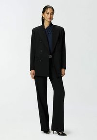 Crni dvoredni sako s reverima, uparen s krojenim crnim hlačama i cipelama s oštrim nosom na visoku petu. Glatka tkanina, strukturirani kroj.