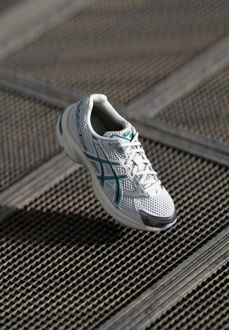 Chaussure de course blanche et bleu canard suspendue au-dessus d’un sol en grille métallique texturée à la lumière naturelle.