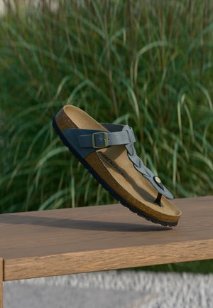 Sininen Birkenstock-sandaali soljella ja varvasremmillä leijuu puupenkin yläpuolella, taustalla pitkää vihreää ruohoa.