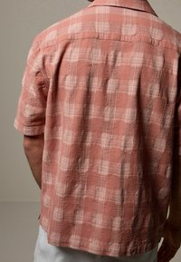Camisa de manga corta en color rosa empolvado, con un patrón de cuadros texturizado, cuello abotonado y un corte relajado con dobladillo recto.