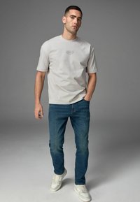 Tricou din bumbac gri deschis, cu mâneci scurte și guler rotund, având un mic grafic imprimat. Purtați-l cu blugi bleu închis și adidași albi.