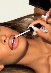 Gloss à lèvres dans un tube noir appliqué sur des lèvres pleines et brillantes. La peau est lisse avec un ton chaud ; les ongles sont longs et peints en blanc. Le fond est uni.