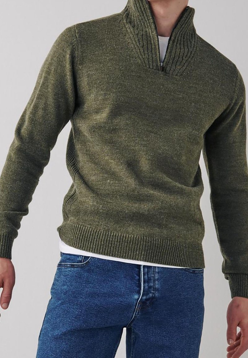 Pull en tricot vert olive avec col demi-zip, poignets et ourlet côtelés, surface texturée, porté sur une chemise blanche, associé à un jean bleu.