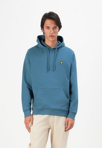 Giovane uomo in piedi che indossa una felpa con cappuccio blu con un emblema giallo e pantaloni beige, davanti a uno sfondo bianco uniforme.