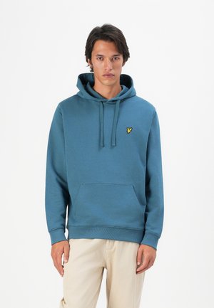 Young man standing wearing a blue hooded sweatshirt with a yellow emblem and beige pants against a plain white background.