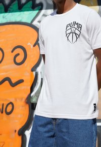 Persona con camiseta blanca de Puma con logo de baloncesto y jeans azules, de pie frente a una colorida pared de graffiti en naranja y verde.