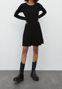 Robe noire côtelée avec des manches longues et une jupe évasée. Le modèle l'associe à des bottes à talons épais noires. Texture lisse et design ajusté.