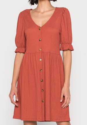 Robe texturée couleur rouille avec boutons à l'avant, col en V, manches courtes bouffantes et poignets à volants portée par une personne debout.