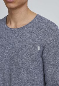 Gray textured knit sweater with a round neckline and a small chest pocket. Features a brand label on the side.