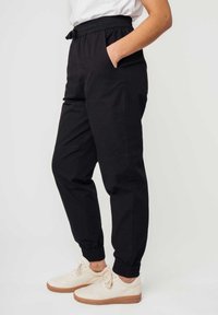 Schwarze Jogginghose mit elastischem Bund, seitlichen Taschen und elastischen Bündchen, aus einem glatten Stoff gefertigt. Getragen mit hellen Sneakers.