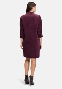 Frau in einem knielangen samtigen Pflaumenkleid mit Dreiviertelärmel und hochhackigen Stiefeletten, die mit dem Rücken zu einer schlichten Hintergrundwand steht.